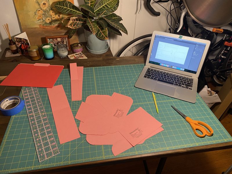 A workbench with pink pattern pieces laid out on a cutting mat, scissors and tools nearby, and a laptop displaying a sewing pattern.